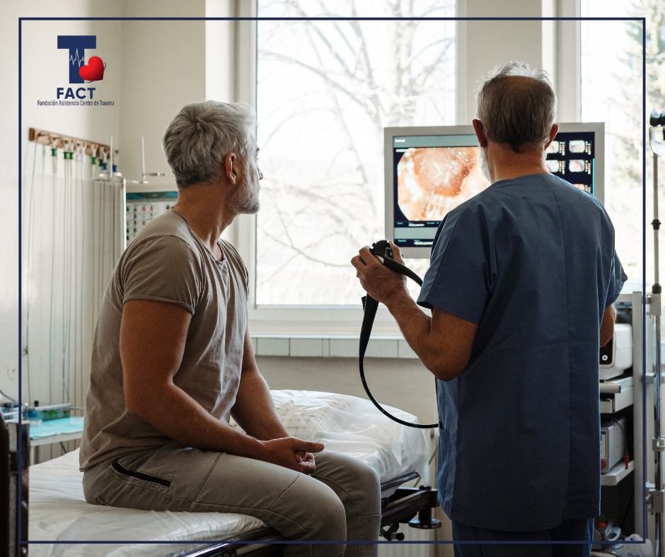Paciente sentado en una camilla mientras un profesional de salud observa un monitor durante un procedimiento diagnóstico en una sala clínica.